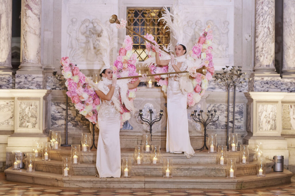 A Vision Of Venice From An International Team Of Creatives Two women with floral wings playing trumpets.