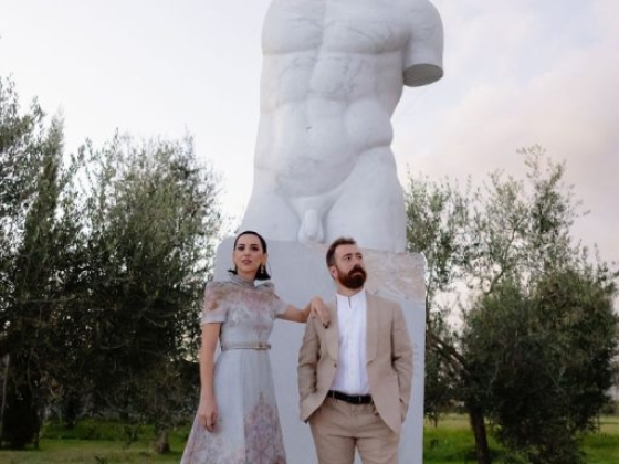 A stylish duo stands near a prominent sculpture.