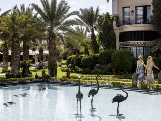 Two women stroll by a decorative pool in a lush garden.