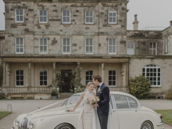Newlyweds pose by a vintage car outside a grand building.