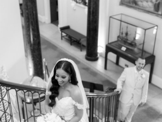 A joyful bride descends stairs, groom looks on.