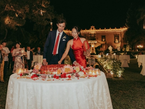 A couple enjoys cake surrounded by guests outdoors.