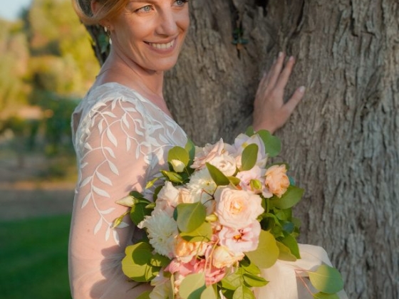 A smiling bride holds a bouquet, standing next to a tree.