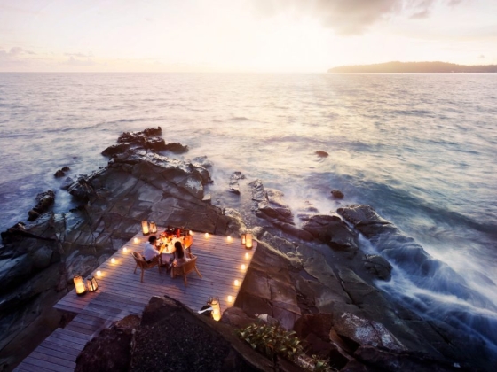 Couple dining by the ocean with lanterns at sunset.