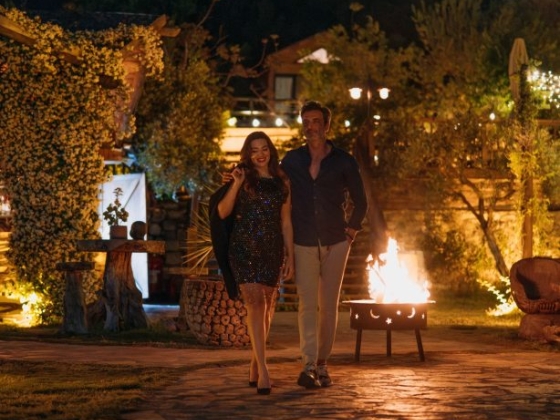 A stylish couple strolls by a warm fire in a garden.