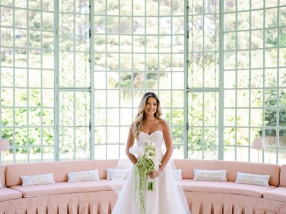 A bride stands elegantly with flowers in a bright, airy space.