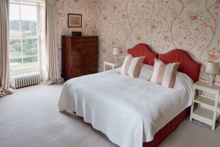 A bedroom with red and white floral wallpaper, a padded headboard and a window over the parkland, Birdsall House