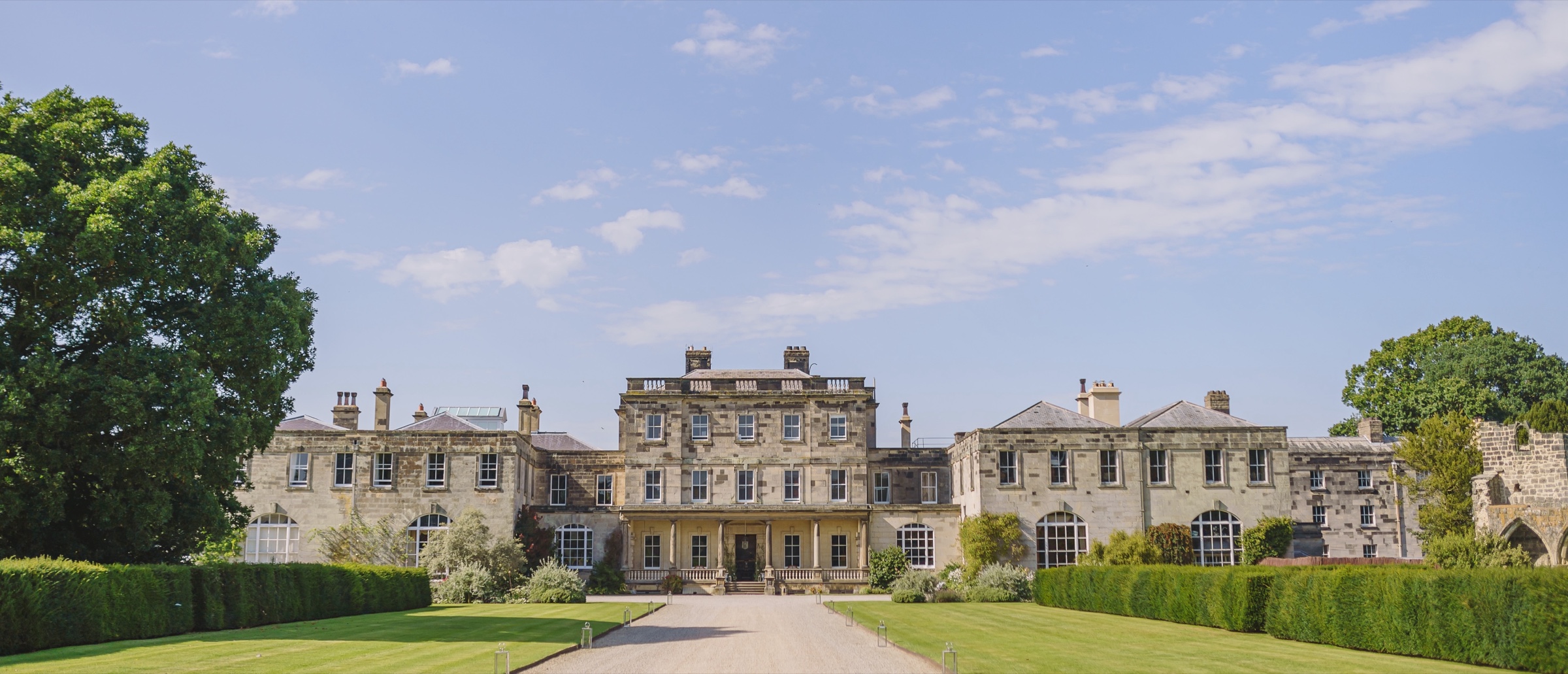 The front of Birdsall House at the end of a gravel drive between clipped hedges and lawns, with the old church ruins at the right edge, Birdsall House
