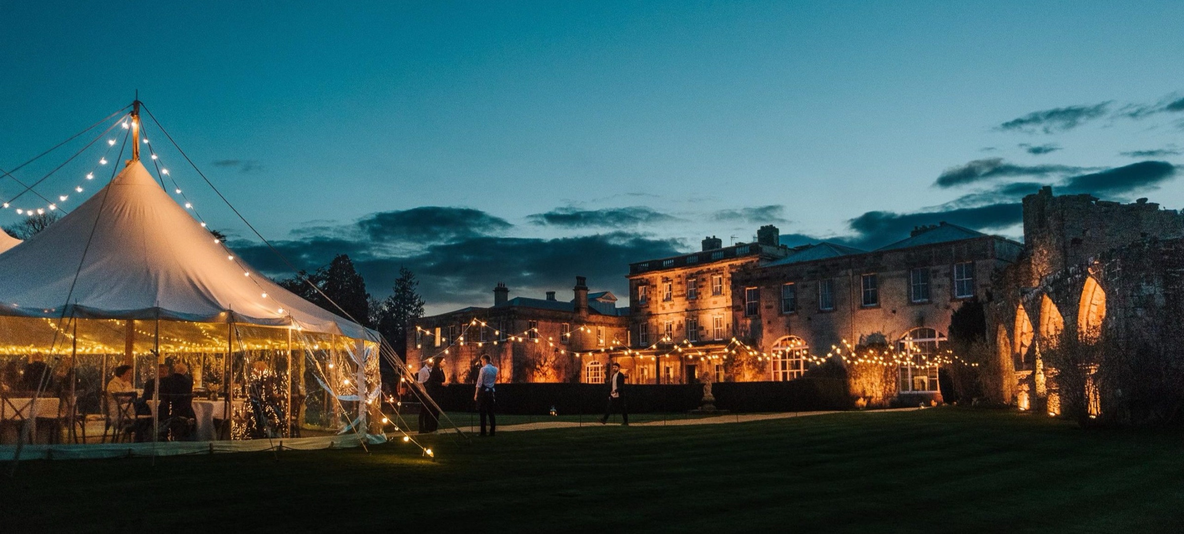 A festoon-lit marquee on the lawn at dusk, with the house and the floodlit arches of the old church ruins behind, Birdsall House