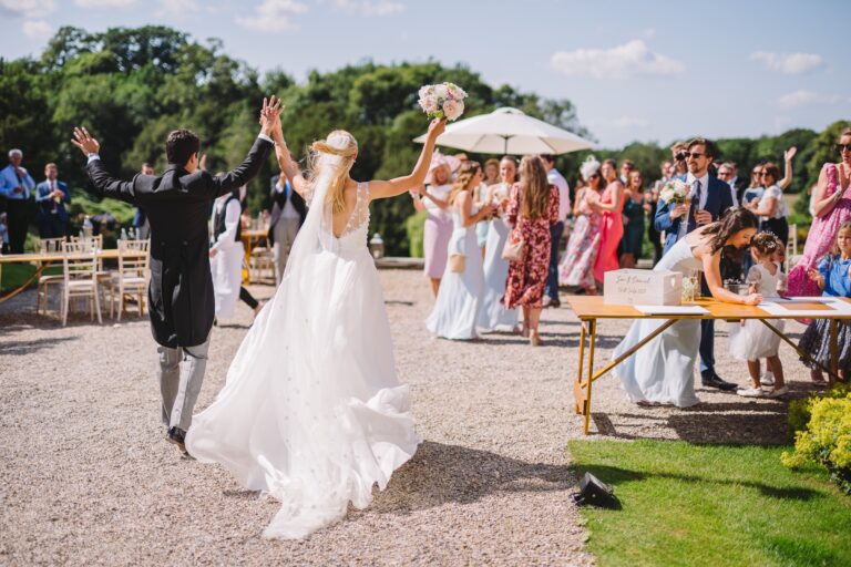 Newlyweds greeting guests on the gravel after the ceremony, with a table for signing and a parasol on the lawn, Birdsall House