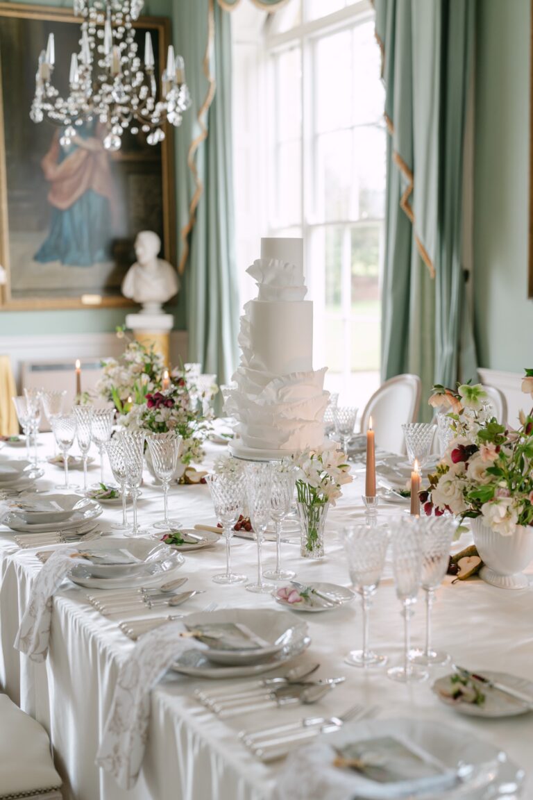 A long table with white linen and a tiered wedding cake beside the tall windows of the Ballroom, Birdsall House