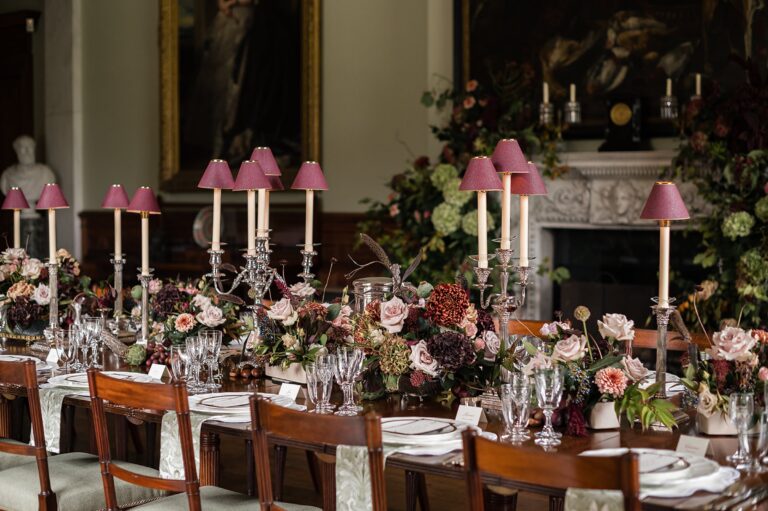 A long dinner table dressed with pink-shaded candle lamps, dark roses and silver candelabra in front of a carved fireplace, Birdsall House