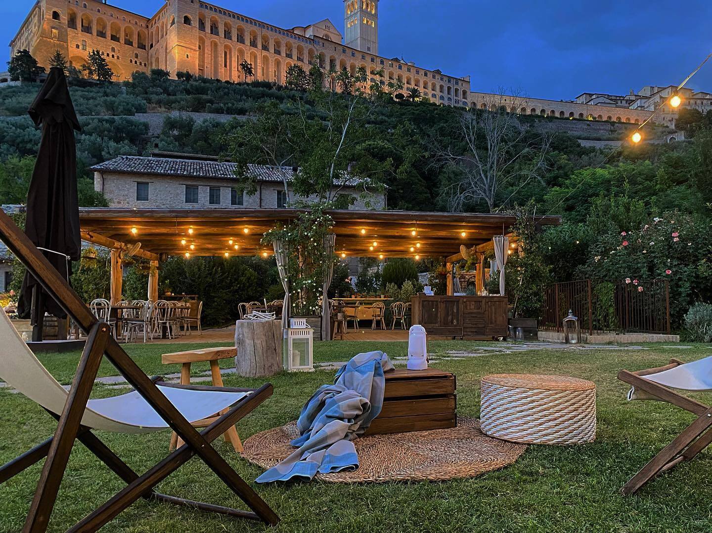The garden at dusk, with deckchairs on the lawn, the lit pergola and the Basilica above, Borgo Antichi Orti Assisi