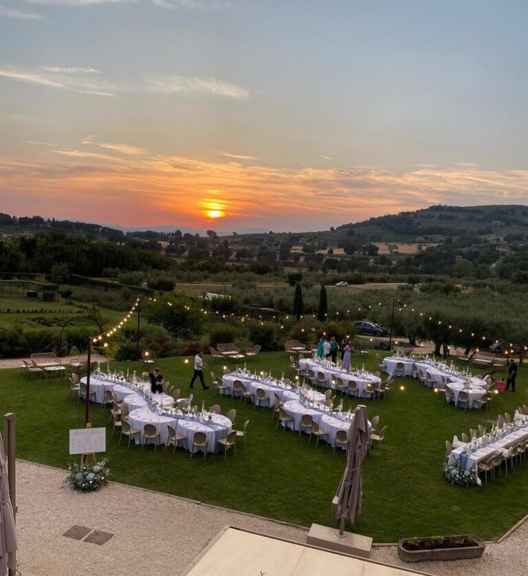Round and long tables laid for a wedding dinner on the lawn, under strings of lights at sunset, Borgo Antichi Orti Assisi