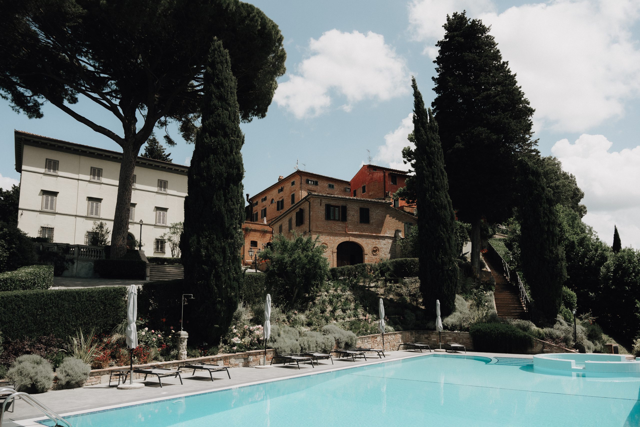 Pool with loungers below the white villa and brick buildings of Borgo Bucciano, framed by cypresses