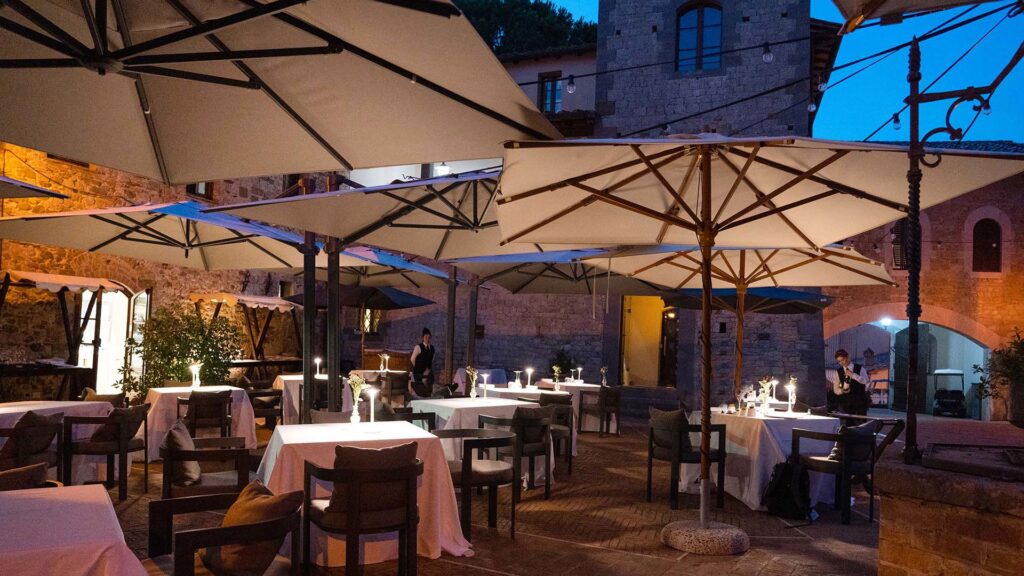 Restaurant tables under umbrellas on the medieval piazza at Castel Monastero at dusk