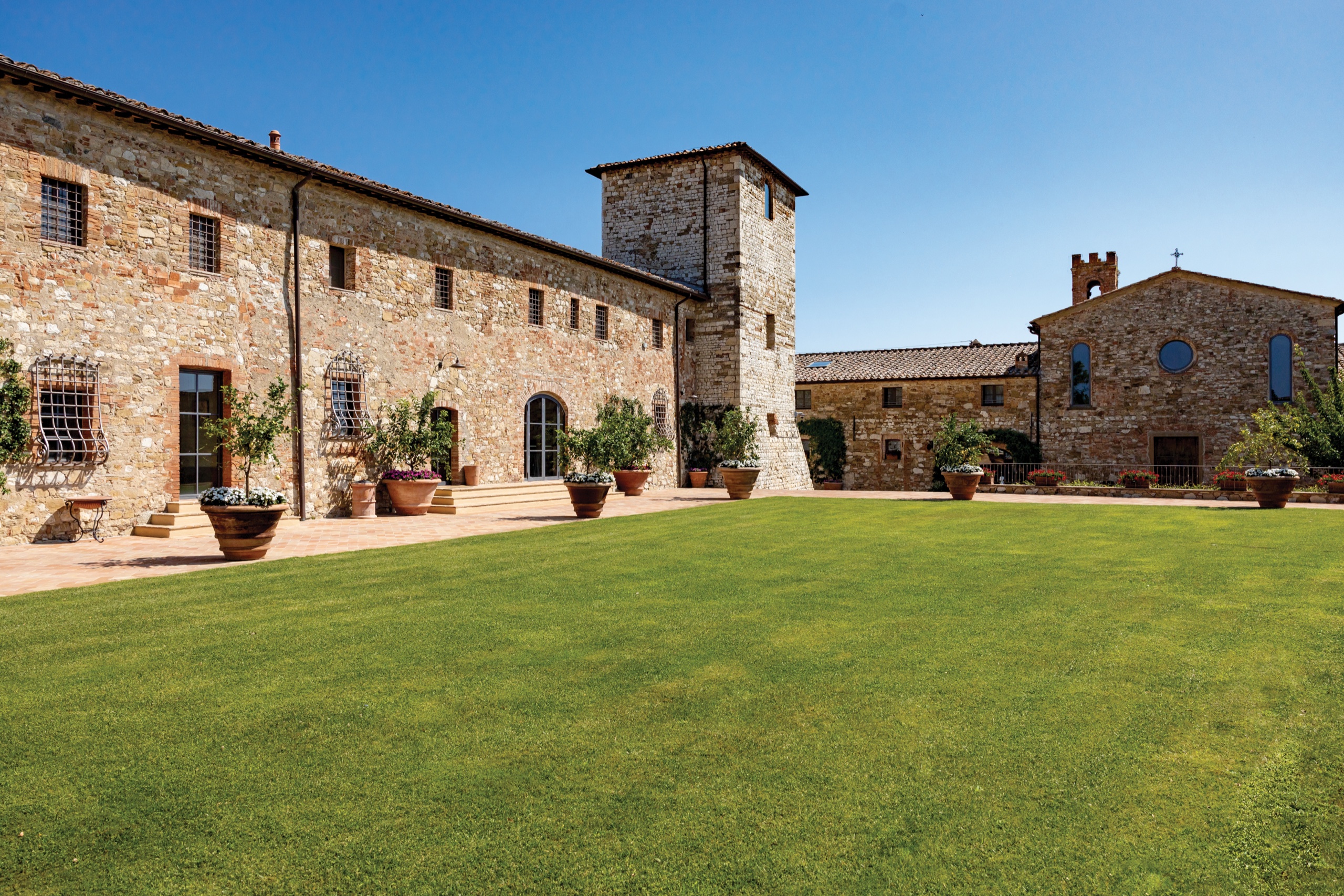 Castle buildings and stone tower facing a broad lawn under blue sky, with a chapel beyond, Castello di Casole