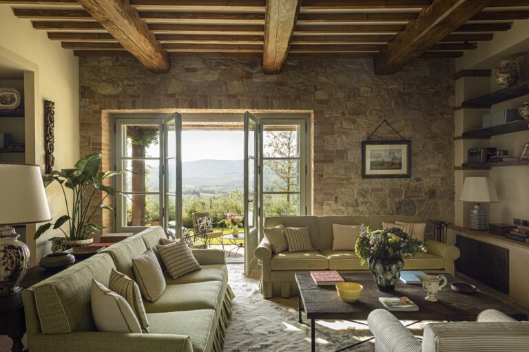 Villa living room with sofas, stone wall, beamed ceiling and glass doors to a hillside view, Castello di Casole