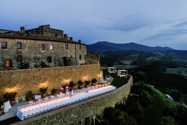 A long candlelit dinner table along the terrace beneath the castle walls at dusk, Castello di Velona Resort