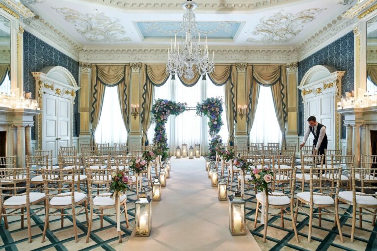 The Drawing Room arranged for a ceremony, with gold chairs, an aisle of lanterns and floral arches by the windows, Claridge's