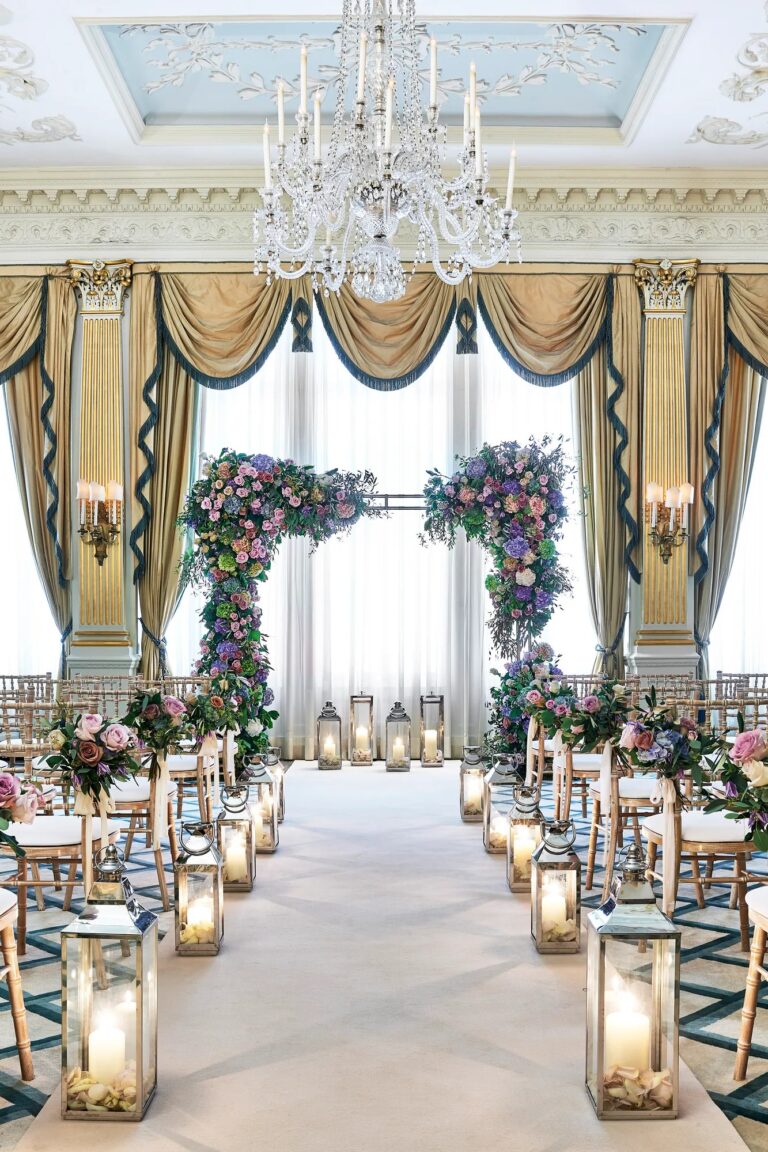 An aisle lined with lanterns and flowers leading to a floral arch in the Drawing Room, Claridge's