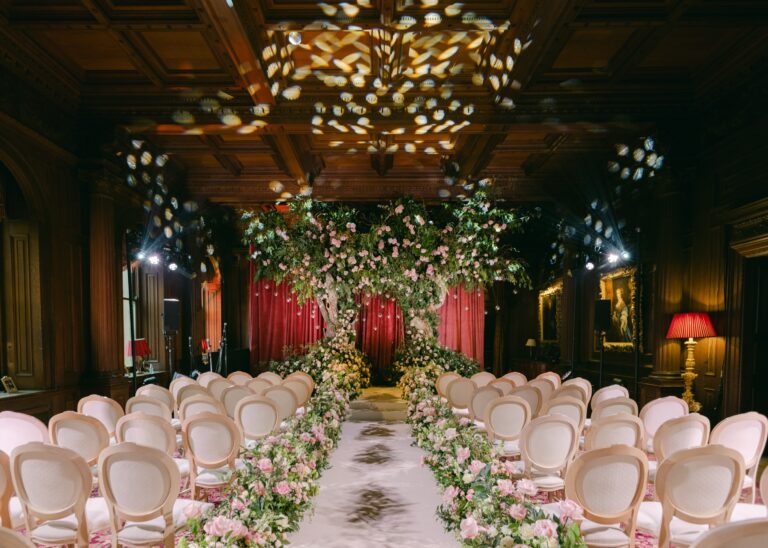 The oak-panelled Great Hall set for a ceremony, with a flowering arch and blush chairs either side of the aisle, Cliveden House