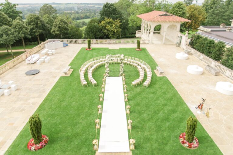 A lawn beside a terracotta-roofed pavilion laid out for an outdoor ceremony, with curved rows of chairs and a white aisle, Cliveden House