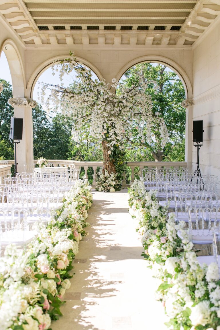 An arched stone loggia with white chairs, a floral aisle and a blossom tree framing views over the trees, Cliveden House