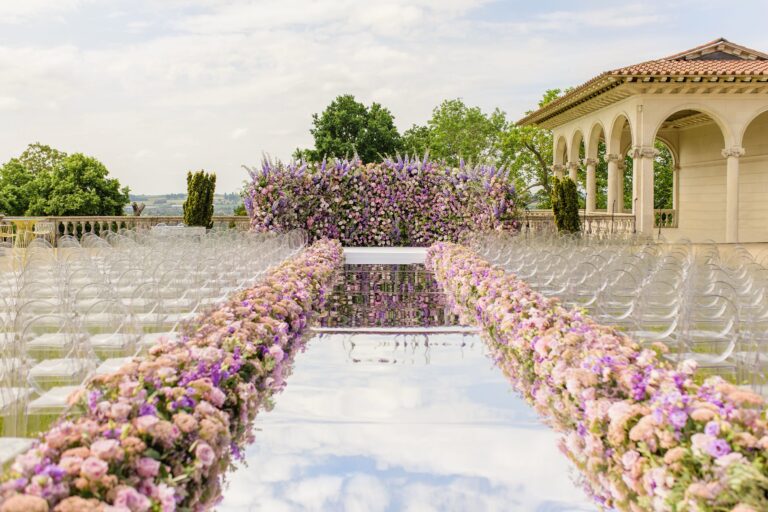 An outdoor ceremony aisle lined with pink and lilac flowers, clear chairs and a wall of blooms beside an arcaded pavilion, Cliveden House