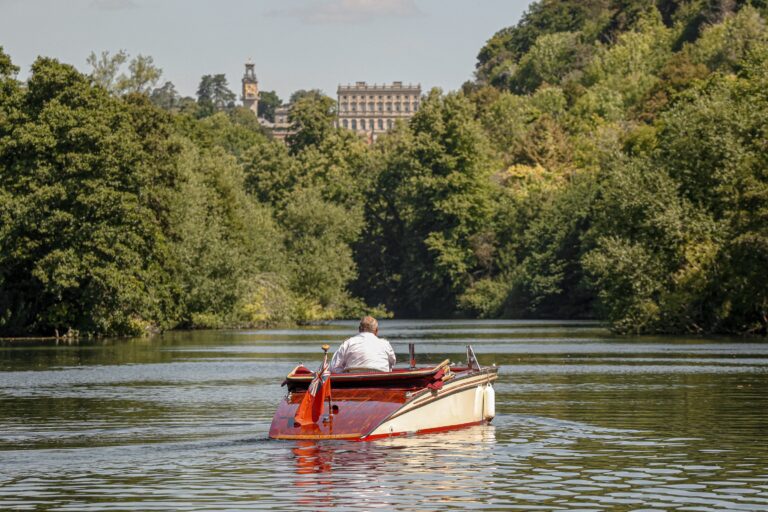 A vintage launch on the Thames below the wooded cliff, with the house visible above the trees, Cliveden House