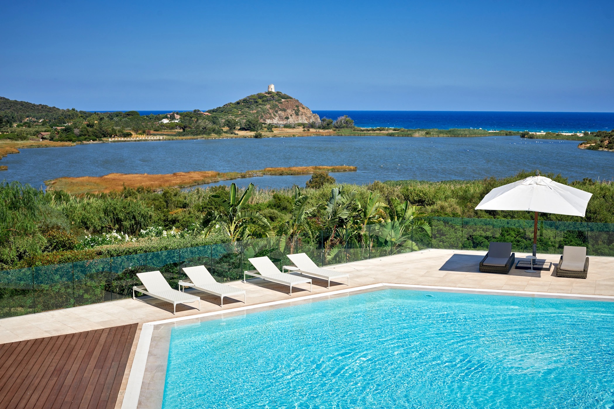 A pool deck with loungers and parasols looking across the lagoon to Chia Tower, Conrad Chia Laguna Sardinia