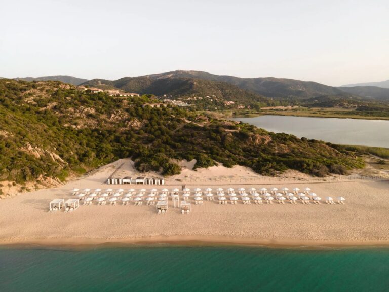 A curve of sand below a terracotta-roofed building, with Chia Tower on its headland across the bay, Conrad Chia Laguna Sardinia