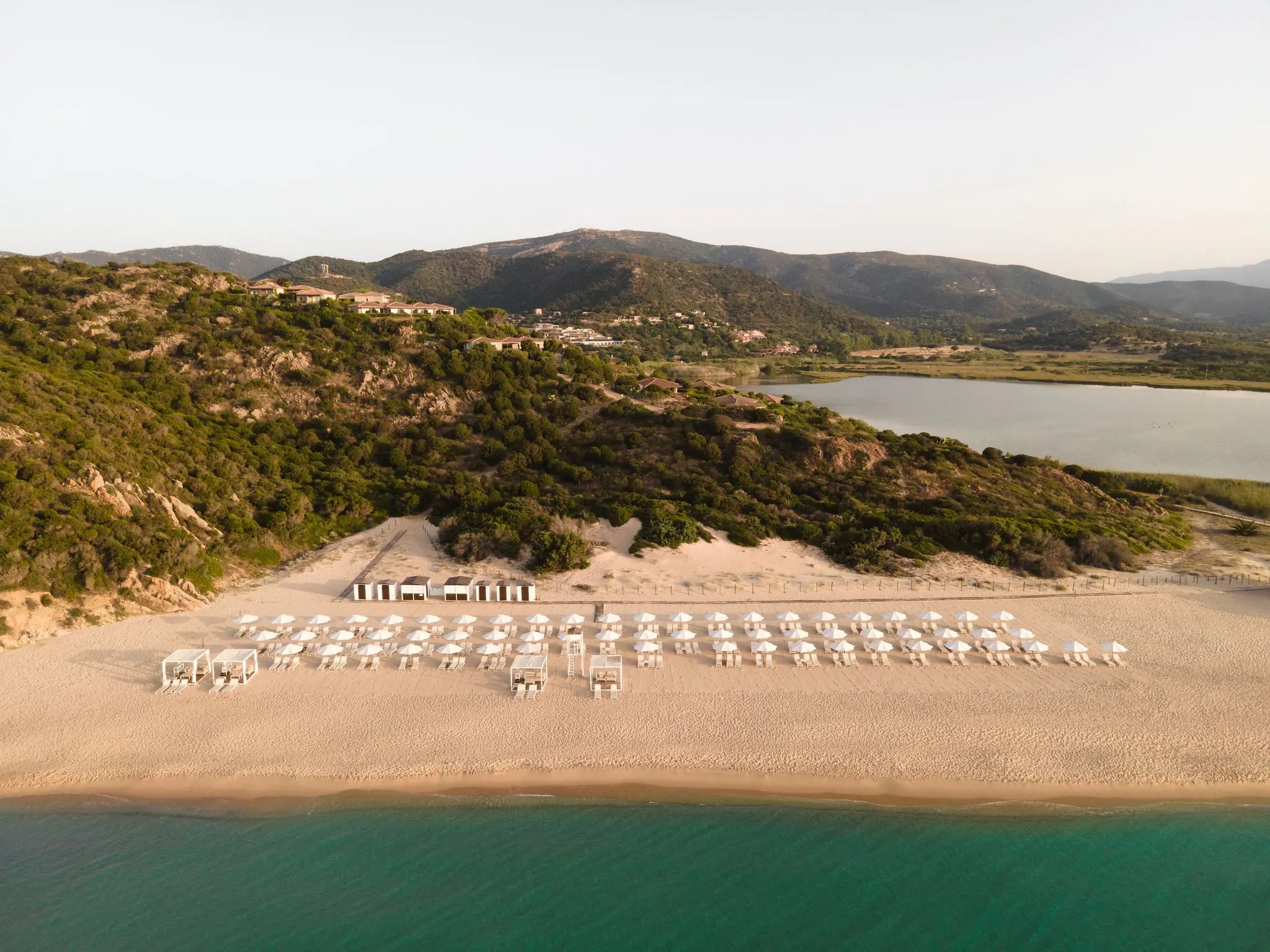 A curve of sand below a terracotta-roofed building, with Chia Tower on its headland across the bay, Conrad Chia Laguna Sardinia