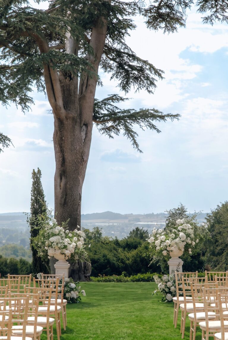 An outdoor ceremony set on the lawn beneath a cedar, with flower urns framing the view over the valley, Hedsor House