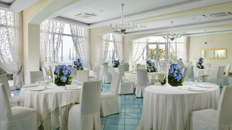 The Ulivi hall laid for a wedding dinner, with blue hydrangeas and sea-facing windows, Hotel Raito