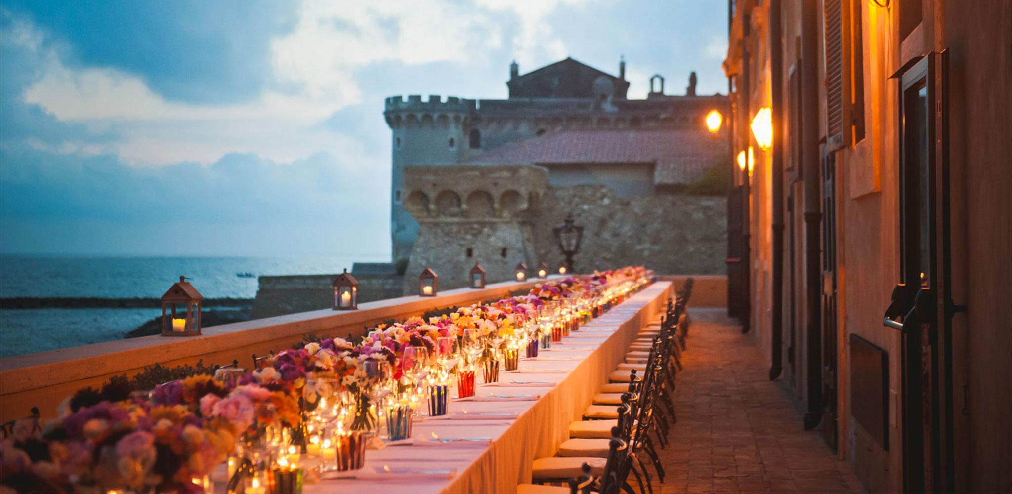 A long candlelit table laid on the sea terrace, with the neighbouring castle behind, La Posta Vecchia Hotel
