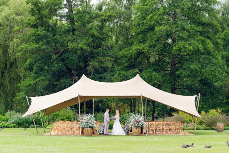 A stretch tent on the lawn under mature trees, set with wooden chairs and flowers for an outdoor ceremony, Leeds Castle