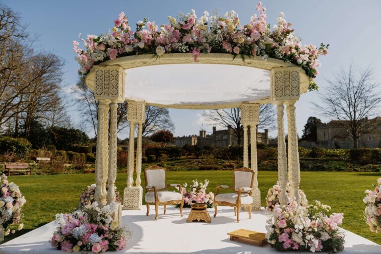 A carved white mandap with pink flowers on the lawn, the Castle visible through the trees behind, Leeds Castle