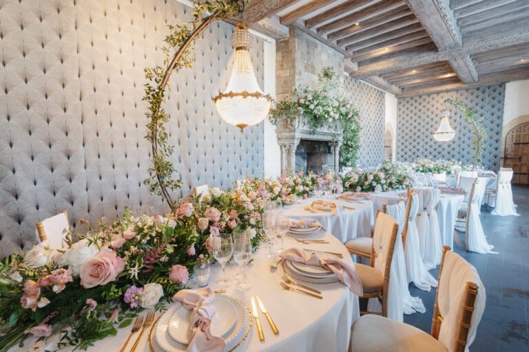 Round tables with pink roses and gold cutlery in the Salon, with a crystal chandelier and the stone fireplace, Leeds Castle