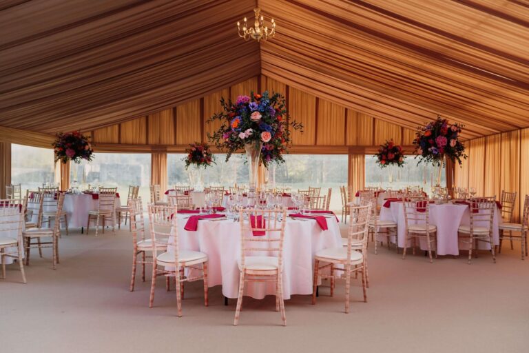 A lined marquee set with round tables, bright centrepieces and a chandelier, with windows onto the grounds, Leeds Castle