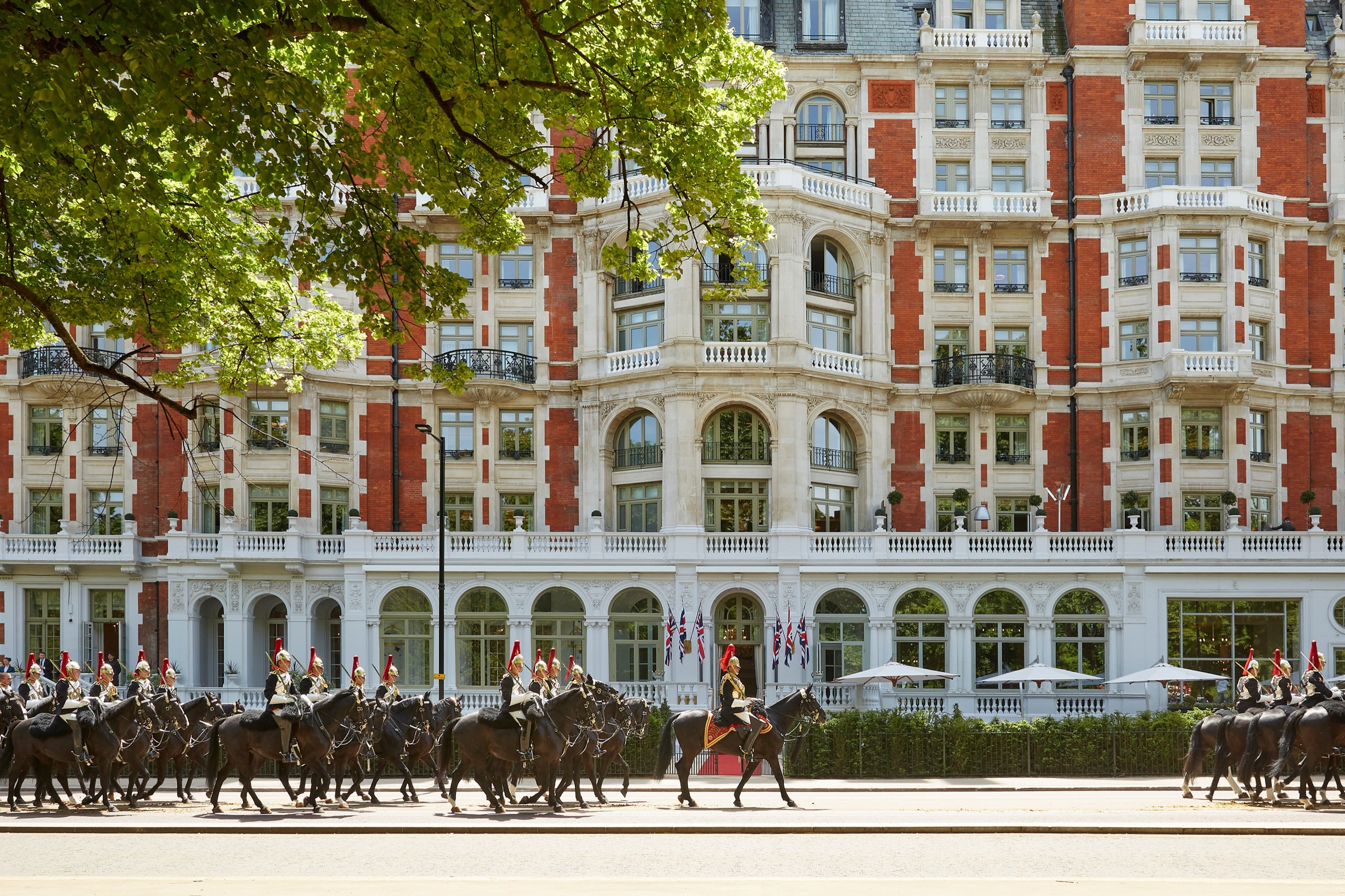 The full park-side facade framed by a tree, with a column of mounted soldiers riding past, Mandarin Oriental Hyde Park