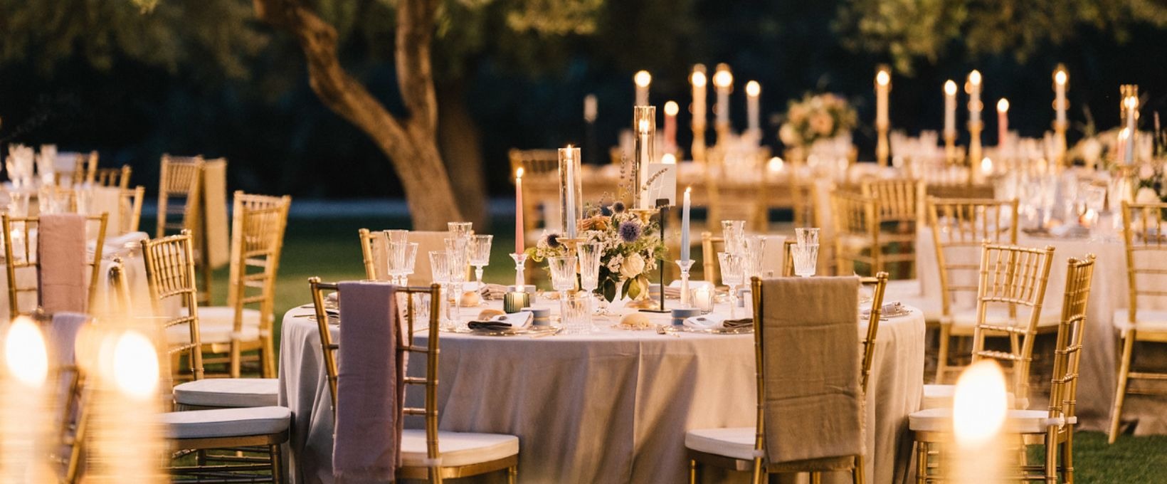 Round tables with gold chairs and tall candles laid for a wedding dinner among the olive trees, Masseria Don Luigi
