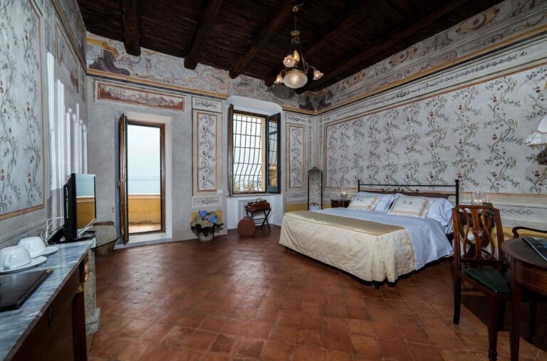 Frescoed bedroom with floral painted walls, beamed ceiling, iron bed and a door open to a sea-view balcony, Palazzo Suriano