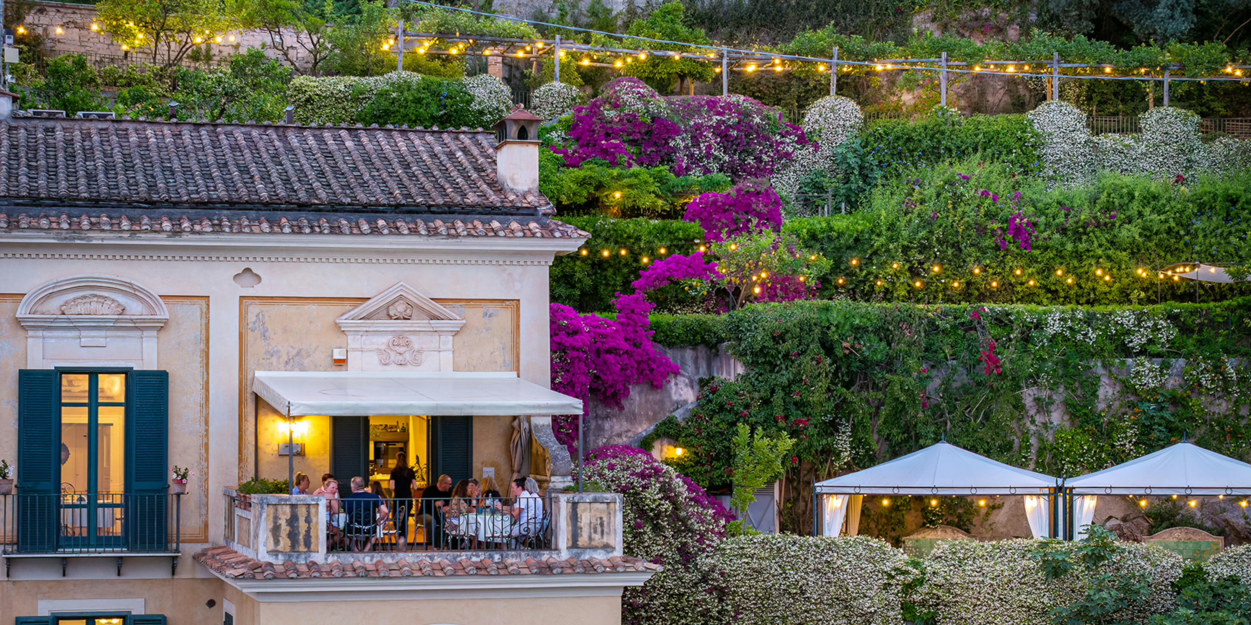 Palazzo terrace lit at dusk with guests dining, terraced gardens with bougainvillea, string lights and white gazebos, Palazzo Suriano