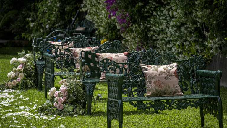 Green cast-iron garden benches with floral cushions and scattered rose petals on the lawn, Palazzo Suriano