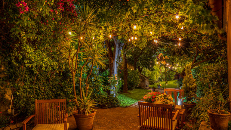 Garden terrace at night with wooden chairs, potted plants and string lights under the trees, Palazzo Suriano