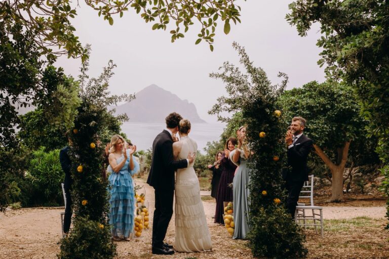 Ceremony under two lemon-hung arches in the garden, guests either side, Monte Cofano in the haze, Pittalumari Makari