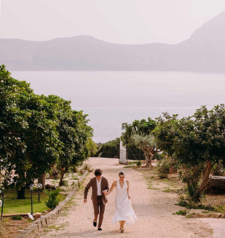 A couple walking up the garden path between trees, the sea and headland behind, Pittalumari Makari