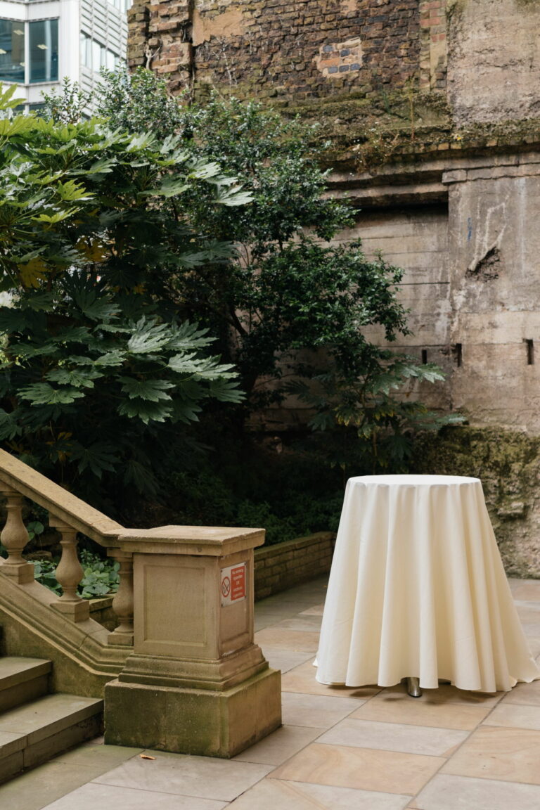 A paved outdoor terrace with stone steps, planting against an old wall and a clothed high table, Plaisterers' Hall