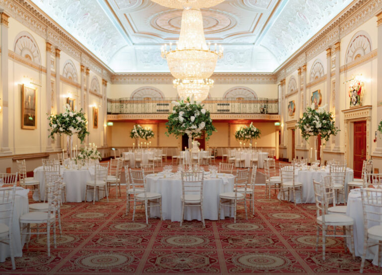 The Great Hall laid with round tables and white chairs under crystal chandeliers, with tall floral centrepieces and a gallery at the far end, Plaisterers' Hall